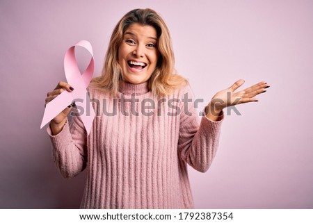 Young beautiful blonde woman asking for support cancer holding pink ribbon symbol very happy and excited, winner expression celebrating victory screaming with big smile and raised hands
