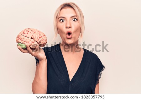 Young beautiful blonde woman asking for care memory holding brain over white background scared and amazed with open mouth for surprise, disbelief face