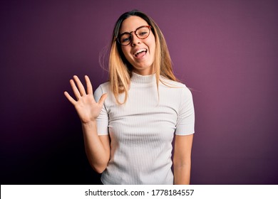 Young beautiful blonde woman with blue eyes wearing casual t-shirt over purple background showing and pointing up with fingers number five while smiling confident and happy.