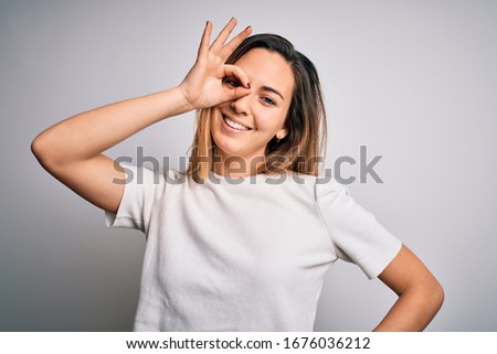 Young beautiful blonde woman with blue eyes wearing casual t-shirt over white background doing ok gesture with hand smiling, eye looking through fingers with happy face.