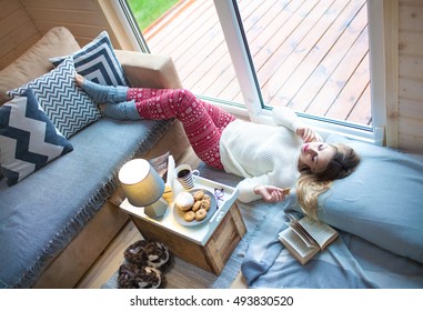 Young beautiful blonde woman with book lying down home by the living room window. Lazy cold day off concept 