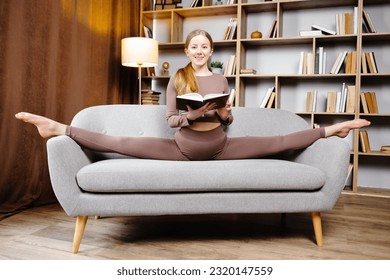 Young beautiful blonde woman in brown leggings and top sitting on a twine at home on the couch. The girl is reading a book sitting on the couch in an unusual position. - Shutterstock ID 2320147559