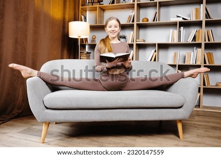 Young beautiful blonde woman in brown leggings and top sitting on a twine at home on the couch. The girl is reading a book sitting on the couch in an unusual position.