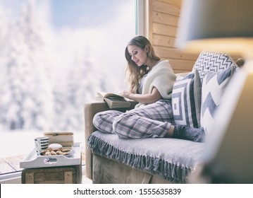 Young Beautiful Blonde Woman With Cup Of Coffee Sitting Home In Living Room By The Window Reading Book. Winter Snow Landscape View. Christmas Concept