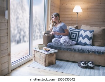 Young Beautiful Blonde Woman With Cup Of Coffee Sitting Home In Living Room By The Window Reading Book. Winter Snow Landscape View. Christmas Concept