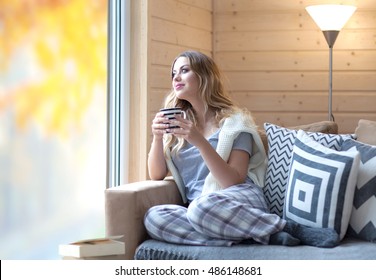 Young beautiful blonde woman with cup of coffee sitting home by the window. Autumn fall trees reflection on the glass. Lazy day off concept