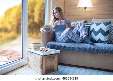 Young Beautiful Blonde Woman With Cup Of Coffee And Book Sitting Home By The Window. Autumn Fall Trees View. Lazy Day Off Concept