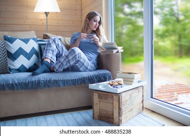 Young Beautiful Blonde Woman With Cup Of Coffee And Book Sitting Home By The Window. Summer Spring Green Woods View. Lazy Day Off Concept