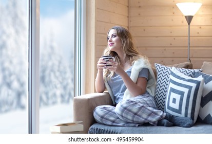 Young beautiful blonde woman with cup of coffee sitting home in living room by the window. Winter snow landscape view. Lazy day off concept