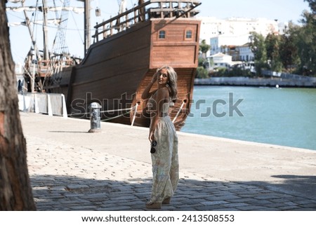 Young beautiful blonde woman dressed in embroidered trousers and crochet top by the river bank in seville. In the background a replica of Christopher Columbus' boat