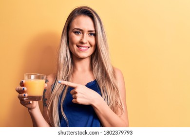 Young beautiful blonde woman drinking glass of healthy orange juice over yellow background smiling happy pointing with hand and finger - Shutterstock ID 1802379793