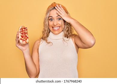 Young beautiful blonde woman eating fast food hot dog with ketchup and mustard stressed and frustrated with hand on head, surprised and angry face