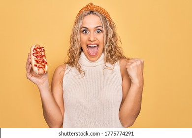 Young beautiful blonde woman eating fast food hot dog with ketchup and mustard screaming proud, celebrating victory and success very excited with raised arms