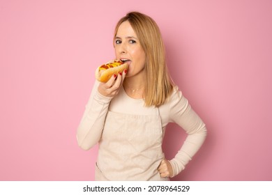 Young Beautiful Blonde Woman Eating Fast Food Hot Dog With Ketchup And Mustard Isolated Over Pink Background.