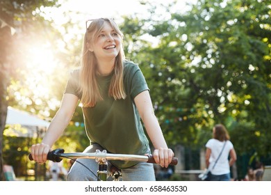 Young Beautiful Blonde Woman Enjoying A Bycicle Ride In The Park During A Food Festival Smiling Off Camera. Copy Space.