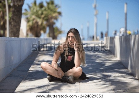 Young, beautiful blonde woman, green eyes, with black top and skirt, tattoos, sitting on the floor, looking at the camera, rebellious and independent. Concept looks, rebelliousness, insubordination.