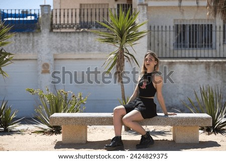 Young, beautiful blonde woman, green eyes, with black top and skirt, tattoos, sitting on a stone bench, looking at the camera rebellious and independent. Concept looks, rebelliousness, insubordination