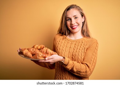 Young beautiful blonde woman holding plate with croissants over isolated yellow background with a happy face standing and smiling with a confident smile showing teeth