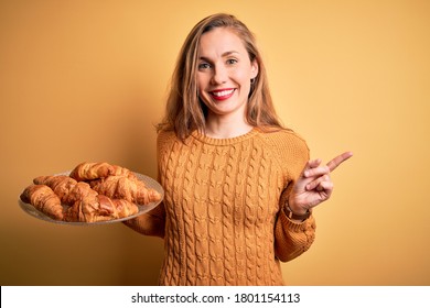 Young beautiful blonde woman holding plate with croissants over isolated yellow background very happy pointing with hand and finger to the side