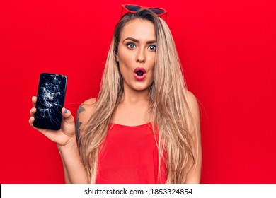 Young beautiful blonde woman holding broken smartphone showing cracked screen scared and amazed with open mouth for surprise, disbelief face