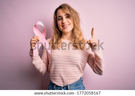 Young beautiful blonde woman holding pink cancer ribbon symbol over isolated background surprised with an idea or question pointing finger with happy face, number one