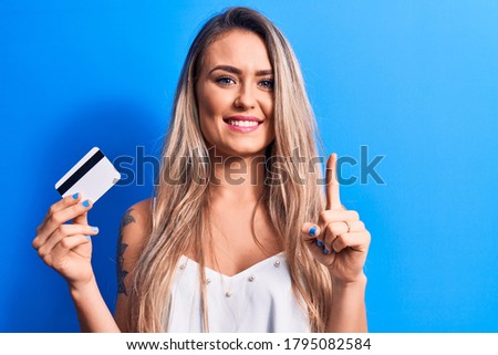 Young beautiful blonde woman holding credit card over isolated blue background smiling with an idea or question pointing finger with happy face, number one