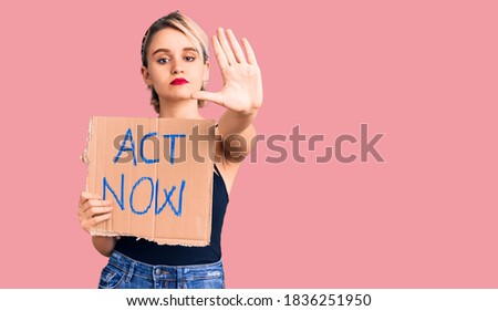 Young beautiful blonde woman holding act now banner with open hand doing stop sign with serious and confident expression, defense gesture 