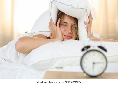 Young Beautiful Blonde Woman Lying In Bed Suffering From Alarm Clock Sound Covering Head And Ears With Pillow Making Unpleasant Face. Early Wake Up, Not Getting Enough Sleep, Going Work Concept