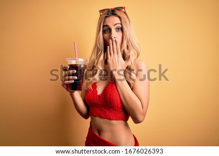 Young beautiful blonde woman on vacation wearing bikini drinking glass of fizzy cola beverage cover mouth with hand shocked with shame for mistake, expression of fear, scared in silence, secret 