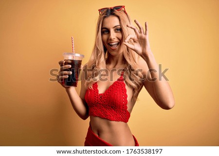 Young beautiful blonde woman on vacation wearing bikini drinking glass of fizzy cola beverage doing ok sign with fingers, excellent symbol