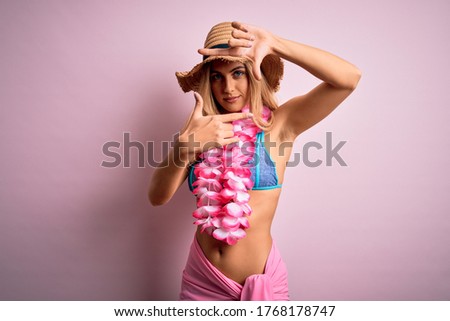 Young beautiful blonde woman on vacation wearing bikini and hat with hawaiian lei flowers smiling making frame with hands and fingers with happy face. Creativity and photography concept.