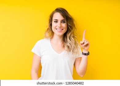 Young beautiful blonde woman over yellow background showing and pointing up with finger number one while smiling confident and happy.