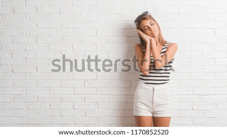 Young beautiful blonde woman over white brick wall sleeping tired dreaming and posing with hands together while smiling with closed eyes.