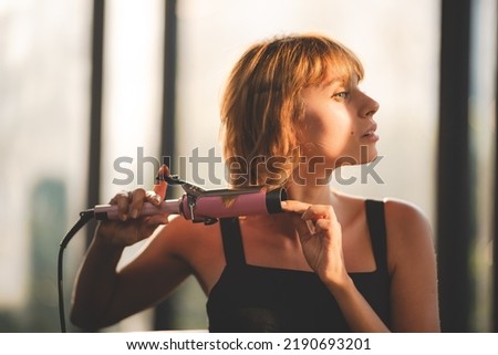 Young beautiful blonde woman with short hair using electronic curling iron styling her hair for fashion and trying new lifestyle at home by looking at self in mirror