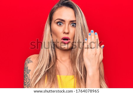 Young beautiful blonde woman showing engagement ring over isolated red background scared and amazed with open mouth for surprise, disbelief face