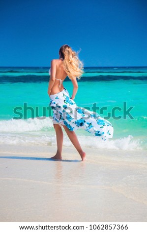 Young beautiful blonde woman with slim tan body in bikini swimwear and long floral skirt on white sand Maldive beach