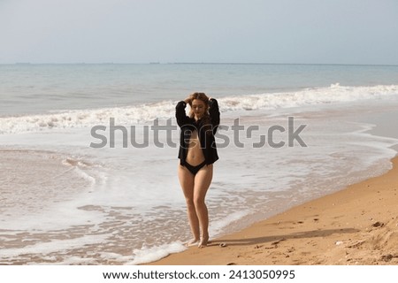 Young and beautiful blonde woman walks along the shore of the beach dressed in black shirt and black bikini and makes different expressions and postures. In the background the sea and the sky.