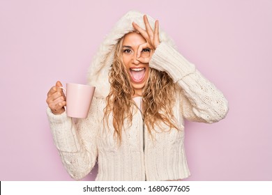 Young beautiful blonde woman wearing casual sweater with hood drinking cup of coffee smiling happy doing ok sign with hand on eye looking through fingers