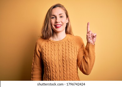 Young beautiful blonde woman wearing casual sweater standing over yellow background showing and pointing up with finger number one while smiling confident and happy.