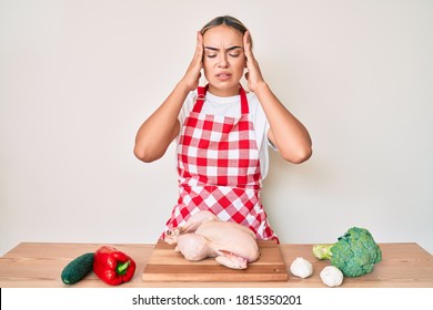 Young Beautiful Blonde Woman Wearing Apron Cooking Chicken With Hand On Head, Headache Because Stress. Suffering Migraine. 