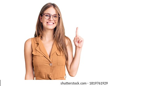 Young beautiful blonde woman wearing casual clothes and glasses showing and pointing up with finger number one while smiling confident and happy. 