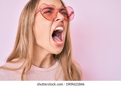 Young beautiful blonde woman wearing heart shaped sunglasses angry and mad screaming frustrated and furious, shouting with anger. rage and aggressive concept.  - Shutterstock ID 1909621846