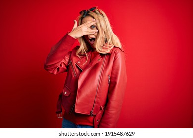 Young Beautiful Blonde Woman Wearing Casual Jacket Standing Over Isolated Red Background Peeking In Shock Covering Face And Eyes With Hand, Looking Through Fingers With Embarrassed Expression.