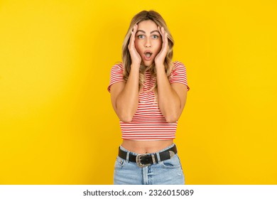 Young beautiful blonde woman wearing striped t-shirt over yellow studio background with scared expression, keeps hands on head, jaw dropped, has terrific expression. Omg concept
