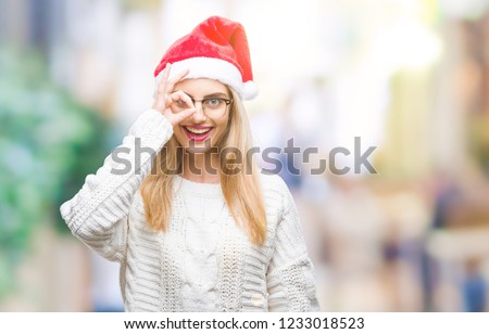 Young beautiful blonde woman wearing christmas hat over isolated background doing ok gesture with hand smiling, eye looking through fingers with happy face.