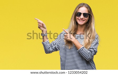 Young beautiful blonde woman wearing sunglasses over isolated background smiling and looking at the camera pointing with two hands and fingers to the side.