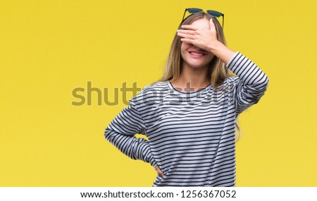 Young beautiful blonde woman wearing sunglasses over isolated background smiling and laughing with hand on face covering eyes for surprise. Blind concept.