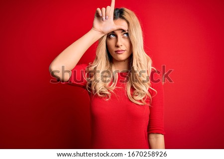 Young beautiful blonde woman wearing casual t-shirt standing over isolated red background making fun of people with fingers on forehead doing loser gesture mocking and insulting.