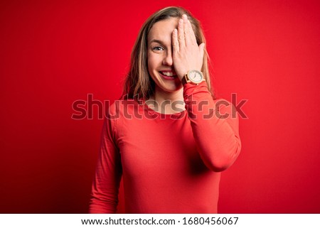 Young beautiful blonde woman wearing casual t-shirt standing over isolated red background covering one eye with hand, confident smile on face and surprise emotion.