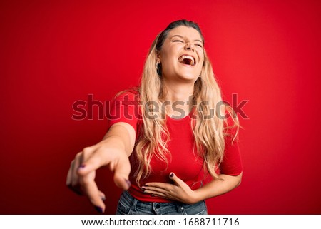 Young beautiful blonde woman wearing casual t-shirt standing over isolated red background laughing at you, pointing finger to the camera with hand over body, shame expression